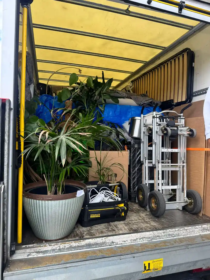 Interior de um caminhão fechado com plantas em vasos, caixa de ferramentas, ferramentas e uma maleta de trabalho, com estrutura metálica e lona azul ao fundo.